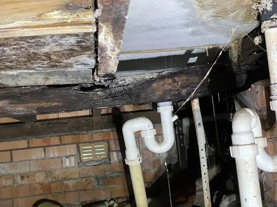 Water-damaged timber beams and PVC pipes beside a brick foundation under a house